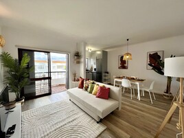 Una sala de estar moderna con un sofá blanco, un comedor con mesa y sillas blancas, y una cocina al fondo.
