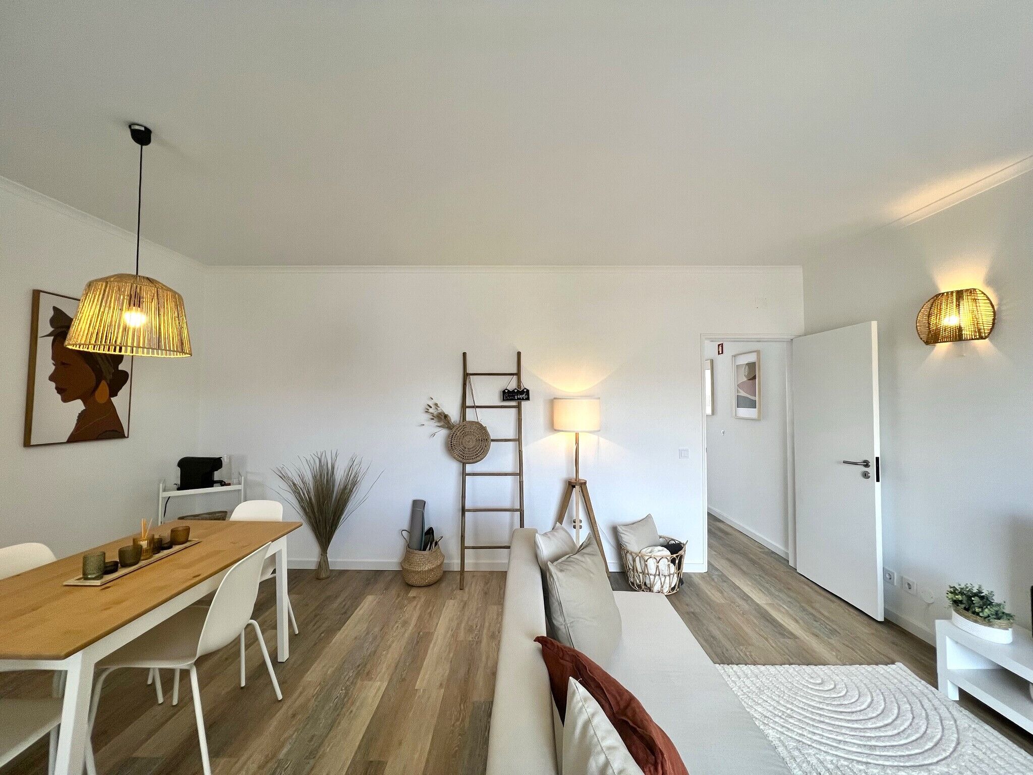A modern living room with a wooden dining table, white chairs, a sofa with cushions, a floor lamp, and a wall-mounted light fixture.