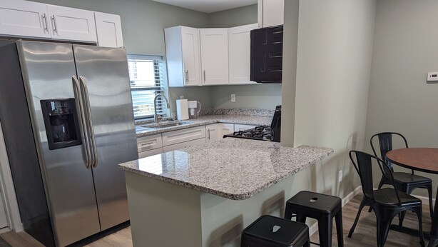 A modern kitchen with a stainless steel refrigerator, granite countertops, white cabinets, a gas stove, and a small dining area with stools. - The Calico Cottage - Fire Pit - Hot Tub (Lewis Center)