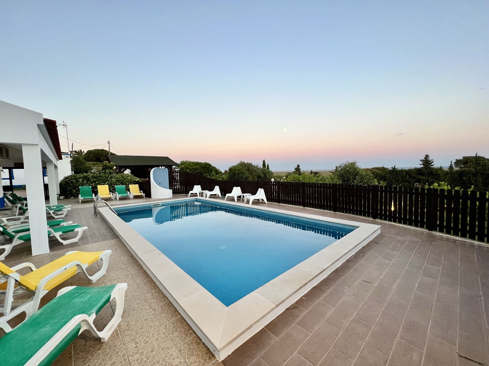 A rectangular outdoor swimming pool with a tiled deck, surrounded by white and green lounge chairs.