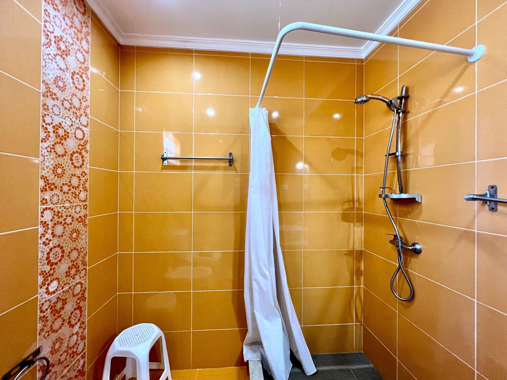 A small bathroom with orange tiles, a shower, a white towel, and a white plastic stool.