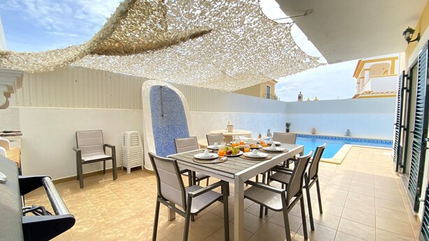 A sunlit patio with a dining table set for two, a pool in the background, and a covered seating area. - Pêra Amazing Villa With Pool by Homing (Silves)