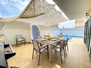 Une terrasse ensoleillée avec une table à manger pour deux, une piscine en arrière-plan et un espace salon abrité.