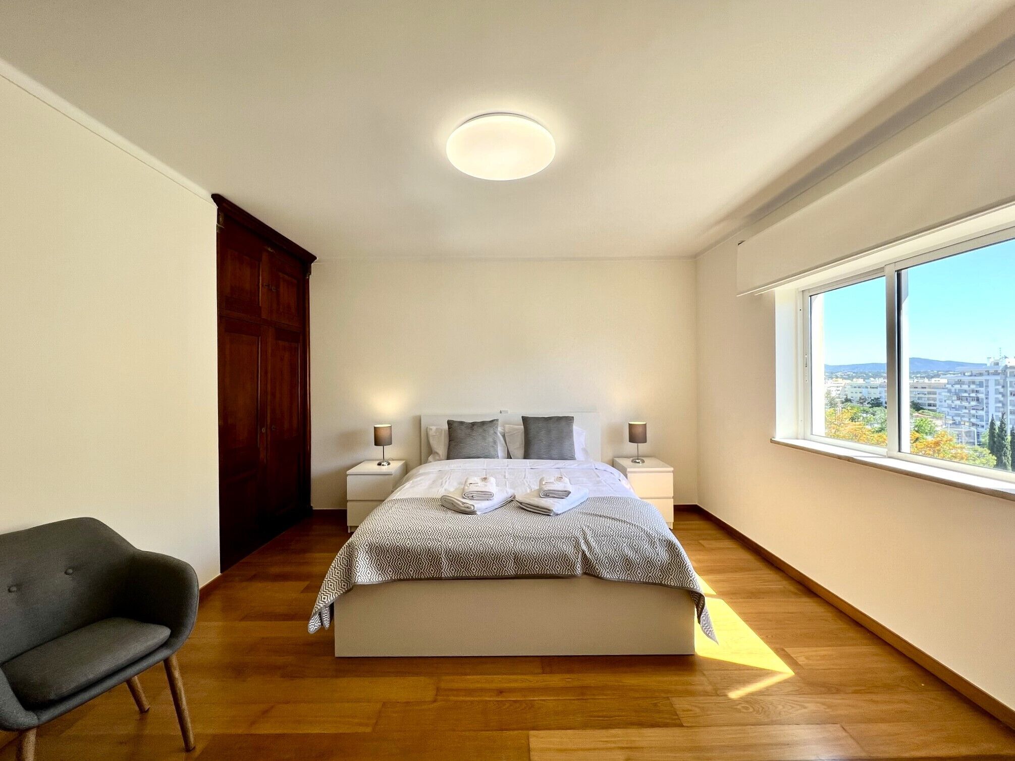 A bedroom with a large bed, two bedside tables with lamps, a wooden wardrobe, a grey armchair, and a large window offering a city view.