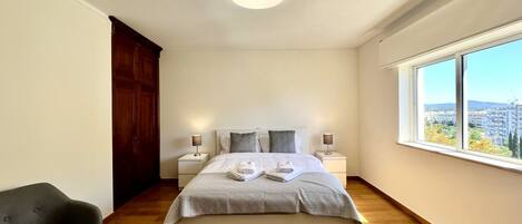 A bedroom with a large bed, two bedside tables with lamps, a wooden wardrobe, a grey armchair, and a large window offering a city view.