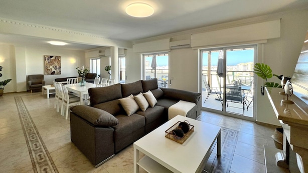 Ein modernes Wohnzimmer mit Sofa, Esstisch und Stühlen. Ein Balkon bietet Blick auf die Stadt.