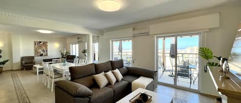 A modern living room with a sofa, dining table, and chairs. There is a balcony with a view of the city.