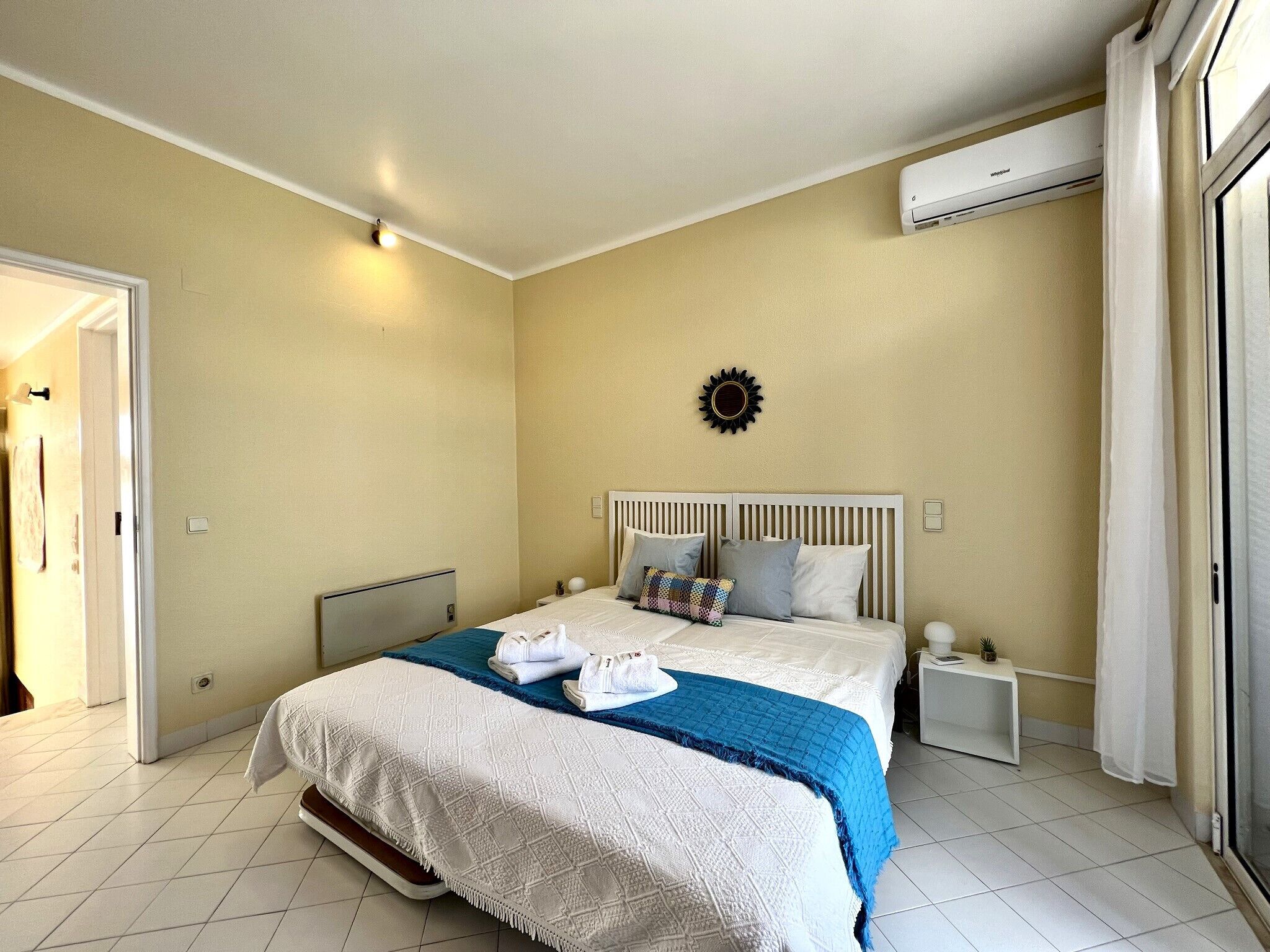 A hotel room with a bed, a nightstand, an air conditioning unit, and a door.