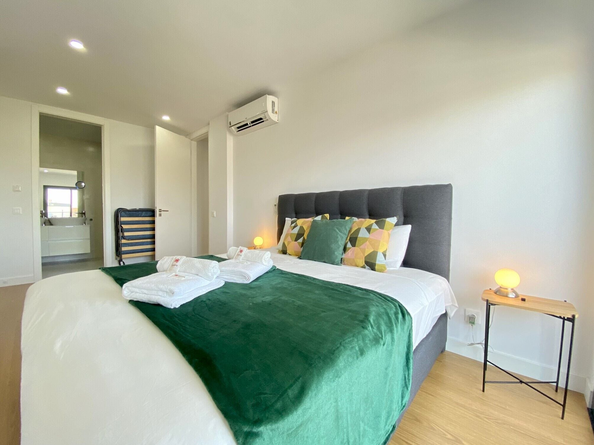 Habitación de hotel con cama, mesita de noche, equipo de aire acondicionado y baño visible a través de una puerta abierta.