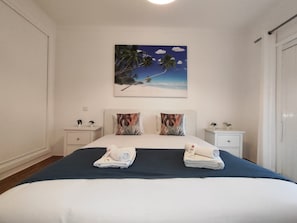 A neatly made bed with two pillows, a nightstand with a plant, and a painting of a beach scene on the wall.