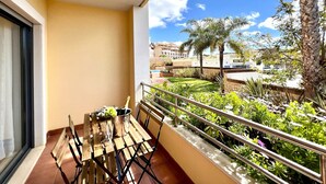 En balkon med et bord, stole og en champagneflaske. Der er gelænder og udsigt til en bygning og grøntområde.