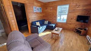 A cozy cabin-style room with a wooden interior, a sofa, a small table, a TV, and a window with a view of trees.