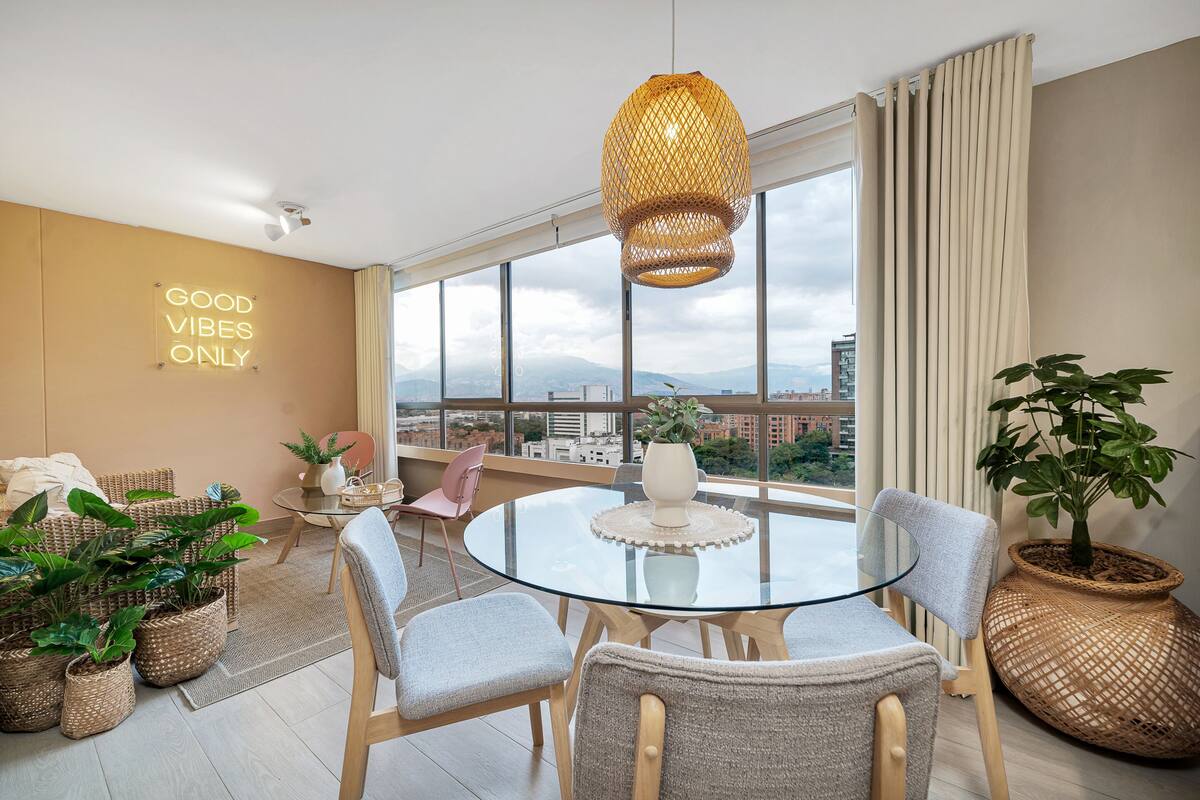 A modern living room with a glass table, wicker chairs, and a large window offering a city view.