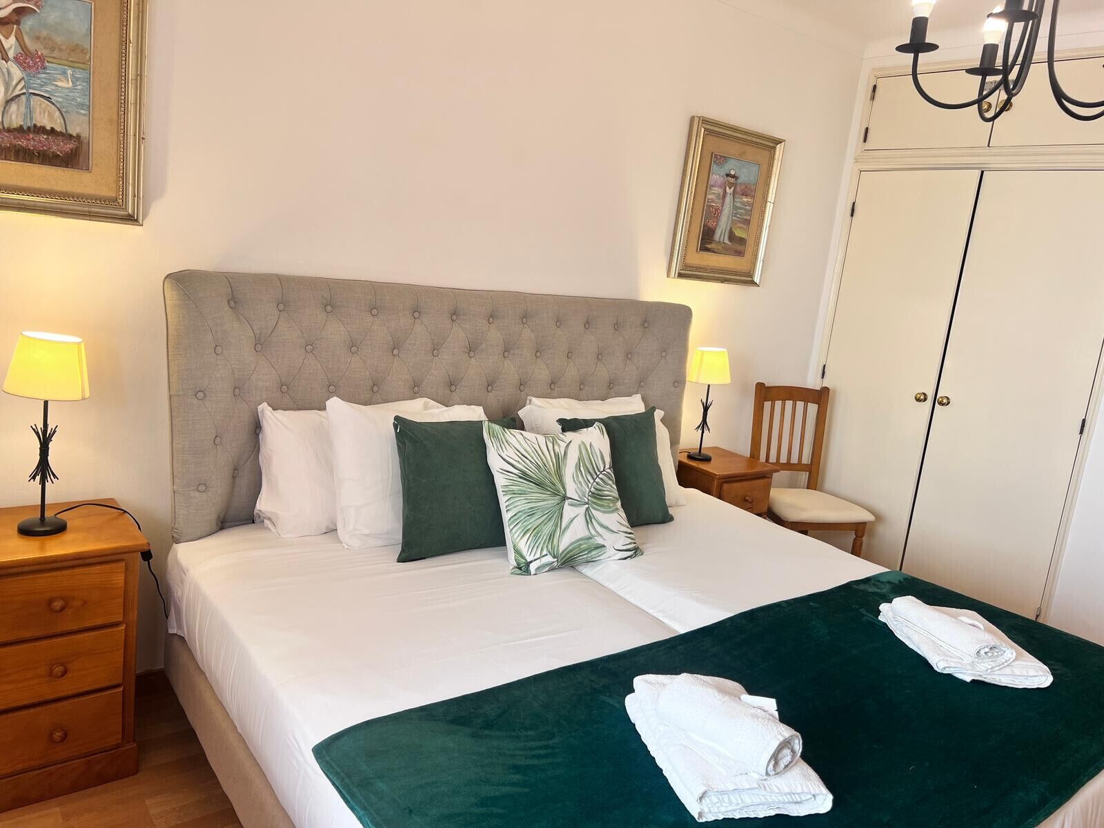 A neatly made bed with a grey upholstered headboard, white and green pillows, and a green throw blanket. There are two bedside tables with lamps, a wooden chair, and a wardrobe with a mirror.