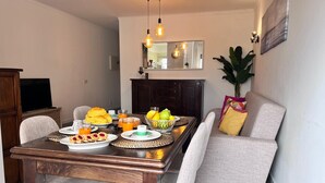 Et spiseområde med et træbord dækket til morgenmad, inklusive croissanter, frugt og drikkevarer. Der er to stole, en sofa med puder og et fjernsyn på et skab.