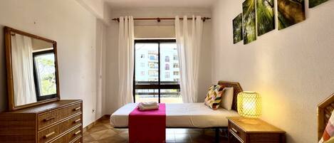 A hotel room with a bed, a dresser, a bedside table with a lamp, a window with curtains, and a view of buildings outside.