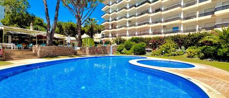 A swimming pool with a stone border, surrounded by a well-maintained garden and a multi-story building with balconies.