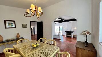 A dining area with a woven table and chairs, a chandelier, and a fireplace in the background.