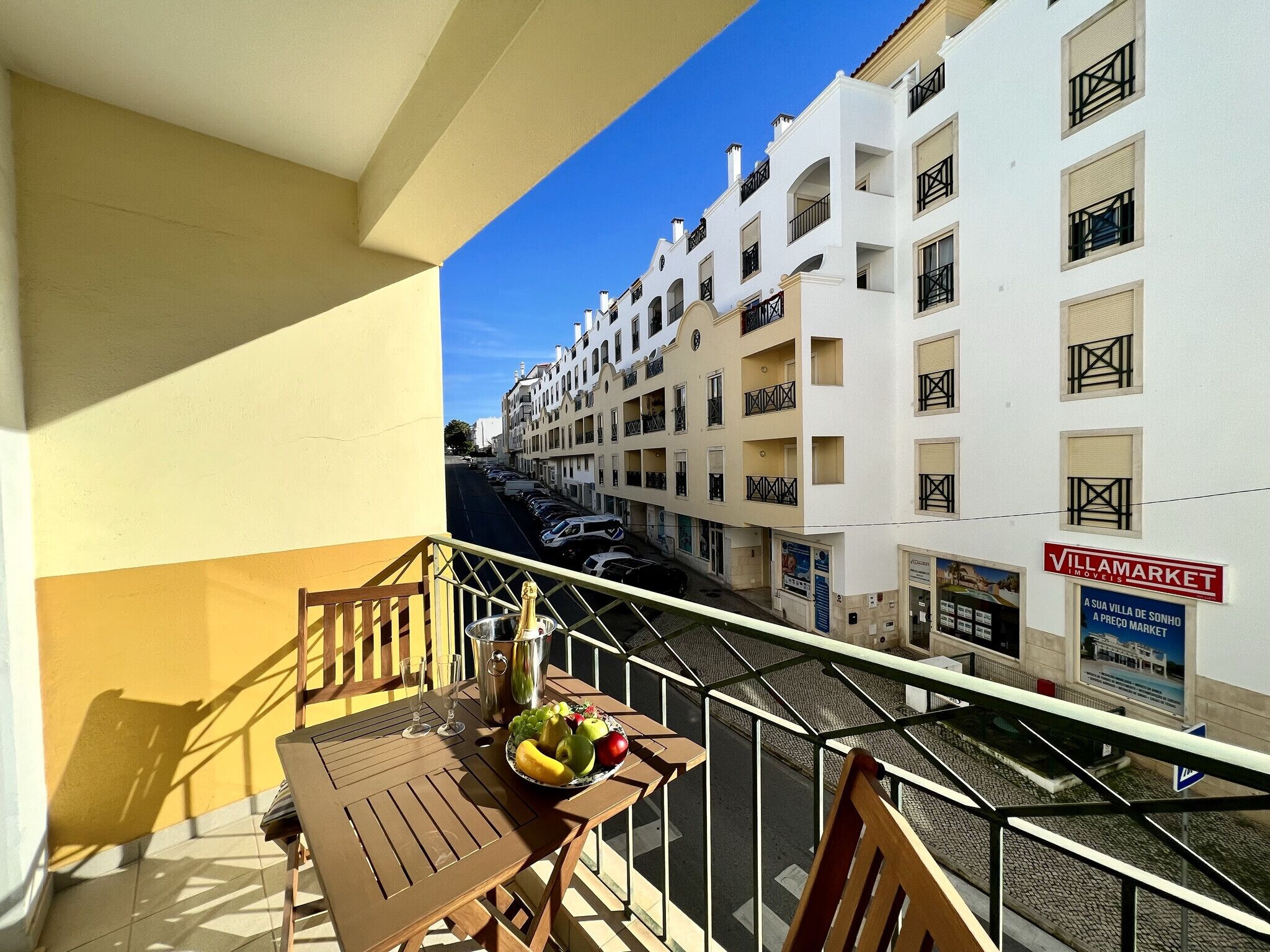 A balcony with a table set for two, a bottle of champagne, and a fruit bowl. The balcony overlooks a street with parked cars and white buildings with balconies.