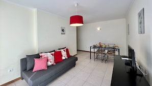 A modern living room with a grey sofa, red and white cushions, a dining table with chairs, a TV on a black stand, and a red pendant light.