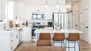 A modern kitchen with white cabinetry, stainless steel appliances, a central island with bar stools, and pendant lighting. - The Table Rocker • Ozark Family Retreat, 5BR 7 BA (Kimberling City)