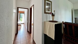 A hallway with a tiled floor, a door leading to a room, a wall-mounted light fixture, and a framed picture above a table with a vase of flowers. - Vilamoura SEA Village by Homing (Loulé)