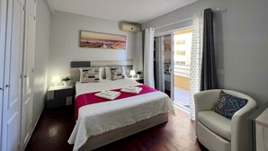 A bedroom with a bed, a chair, a nightstand, a lamp, a painting, and a view of the outside. - Vilamoura Marina MAR 2 With Pool (Loulé)
