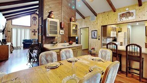 Un grand espace à manger avec une table en bois dressée pour six personnes, entourée de chaises à motifs. On aperçoit, au fond, une cuisine avec des meubles en bois et un réfrigérateur.