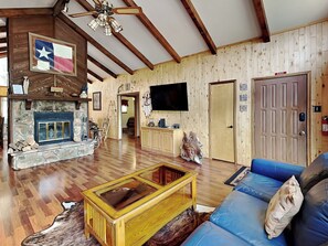 A spacious living room with a stone fireplace, wooden walls, a television, and a blue sofa. - Indianhead Lodge by Ruidoso Vacation Rentals (Ruidoso)