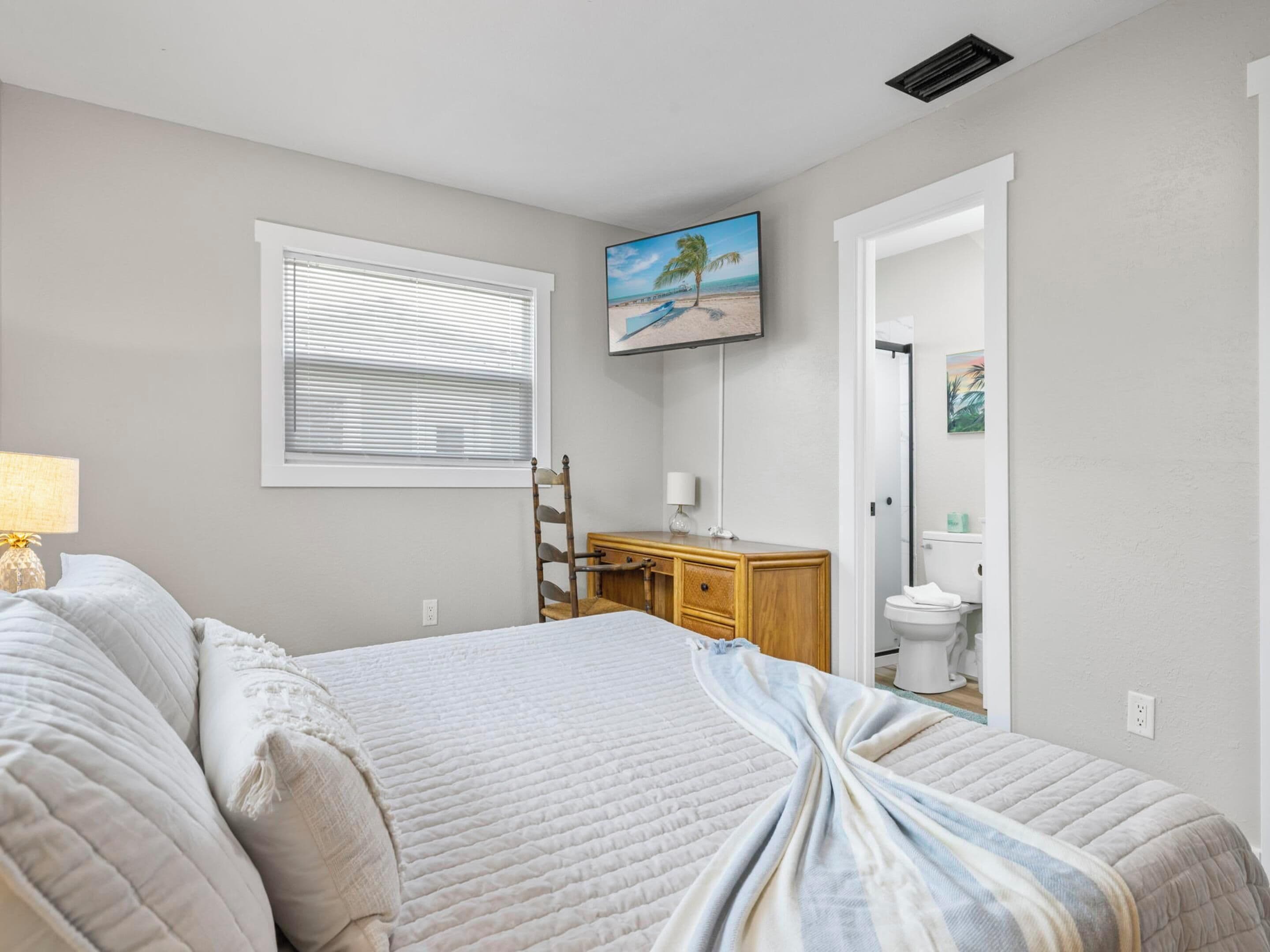 A bedroom with a bed, a bedside table, a chair, a window with blinds, a painting on the wall, and a bathroom visible through an open door.