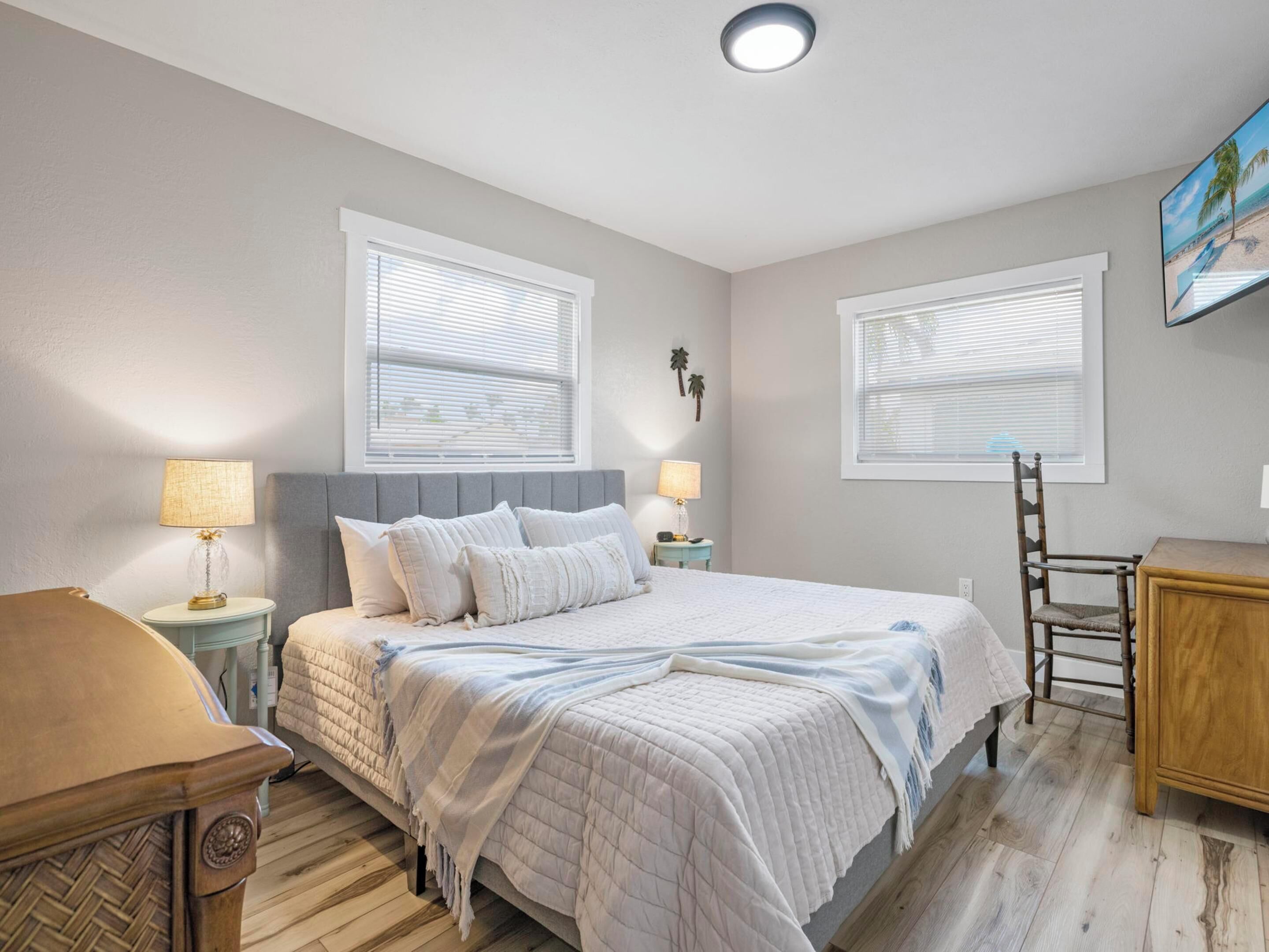 A bedroom with a bed, two bedside tables, a chair, a wooden cabinet, and two windows with blinds.