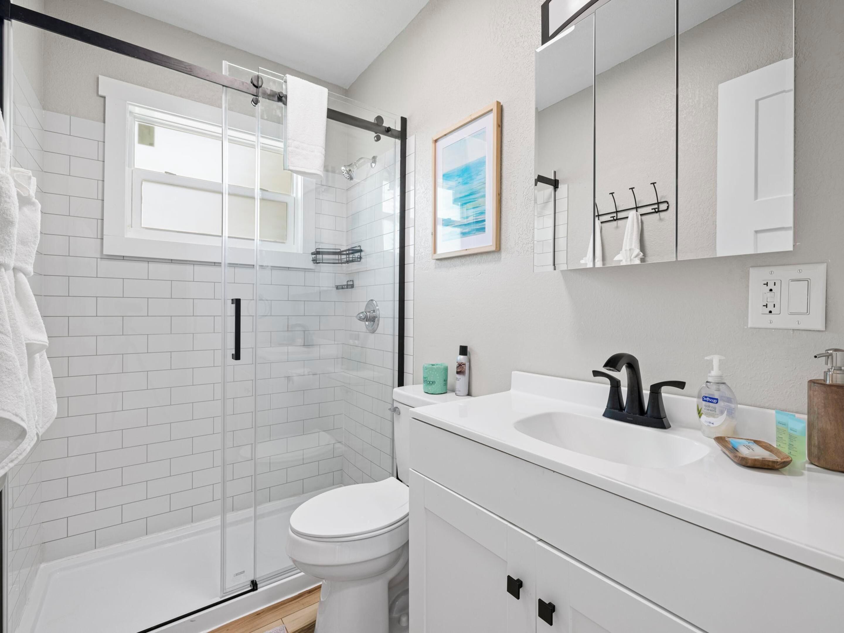 Een moderne badkamer met een glazen douchecabine, witte tegels, een wit toilet, een wastafel met een zwarte kraan en een spiegel boven de wastafel.