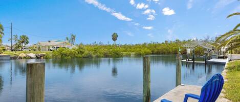 A wooden dock extending into calm water with a blue chair on it, surrounded by greenery and a clear sky.