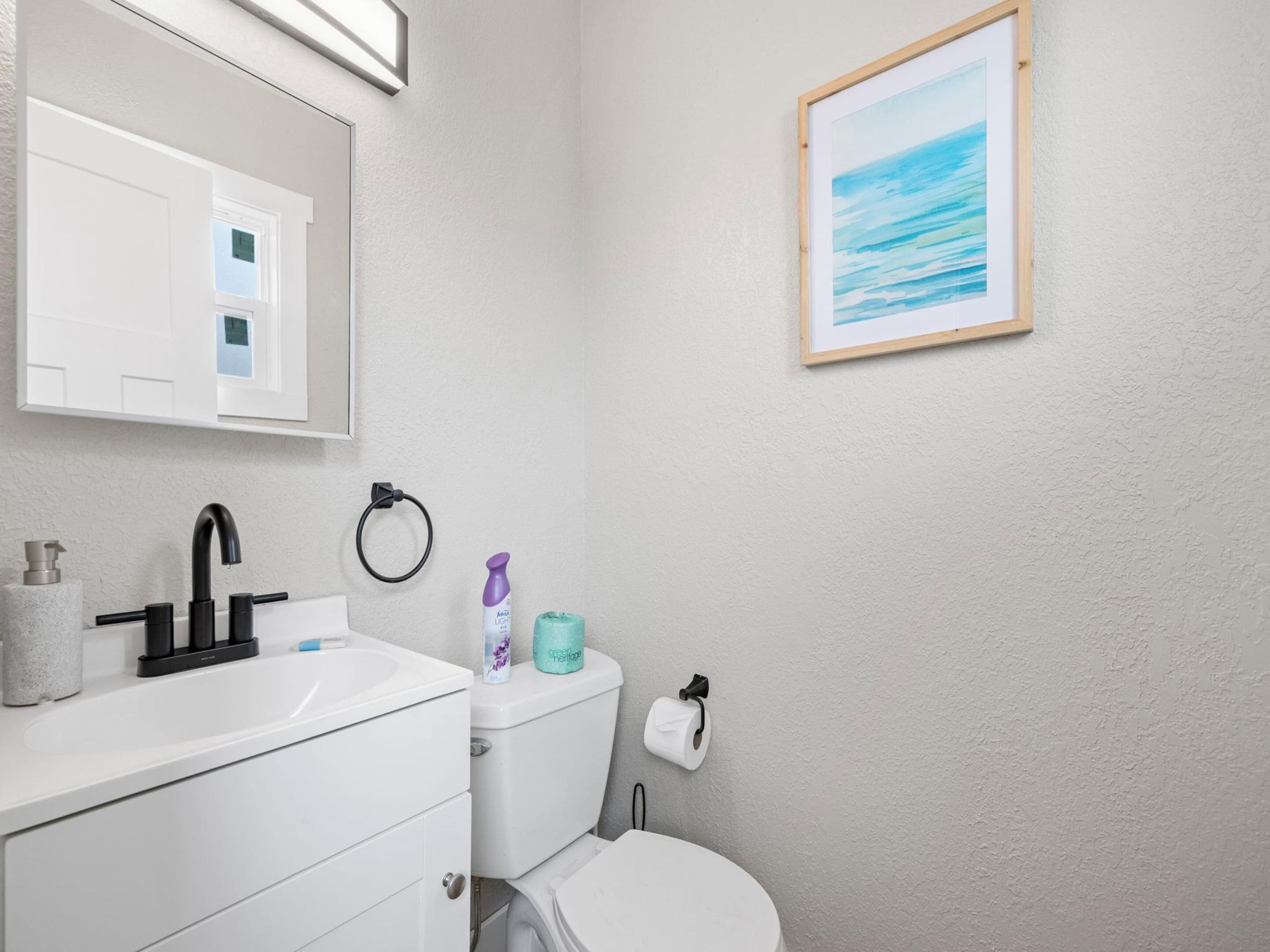 A bathroom with a white sink, black faucet, toilet, and a framed picture of a beach on the wall.