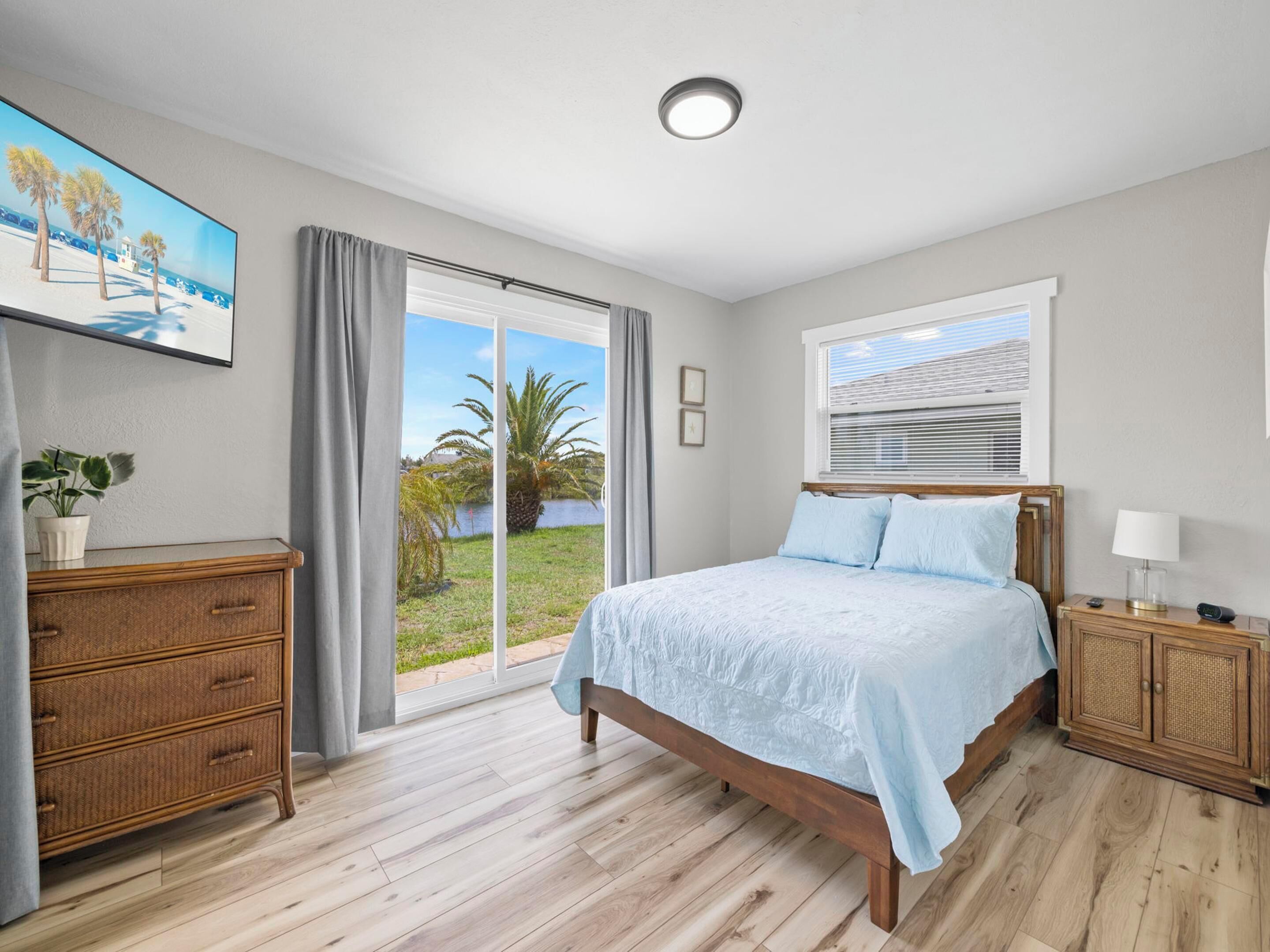 Un dormitorio con cama, mesita de noche, cómoda, ventana con persianas y vista a una palmera y la playa a través de la ventana.