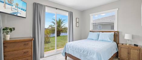 Un dormitorio con cama, mesita de noche, cómoda, ventana con persianas y vista a una palmera y la playa a través de la ventana.