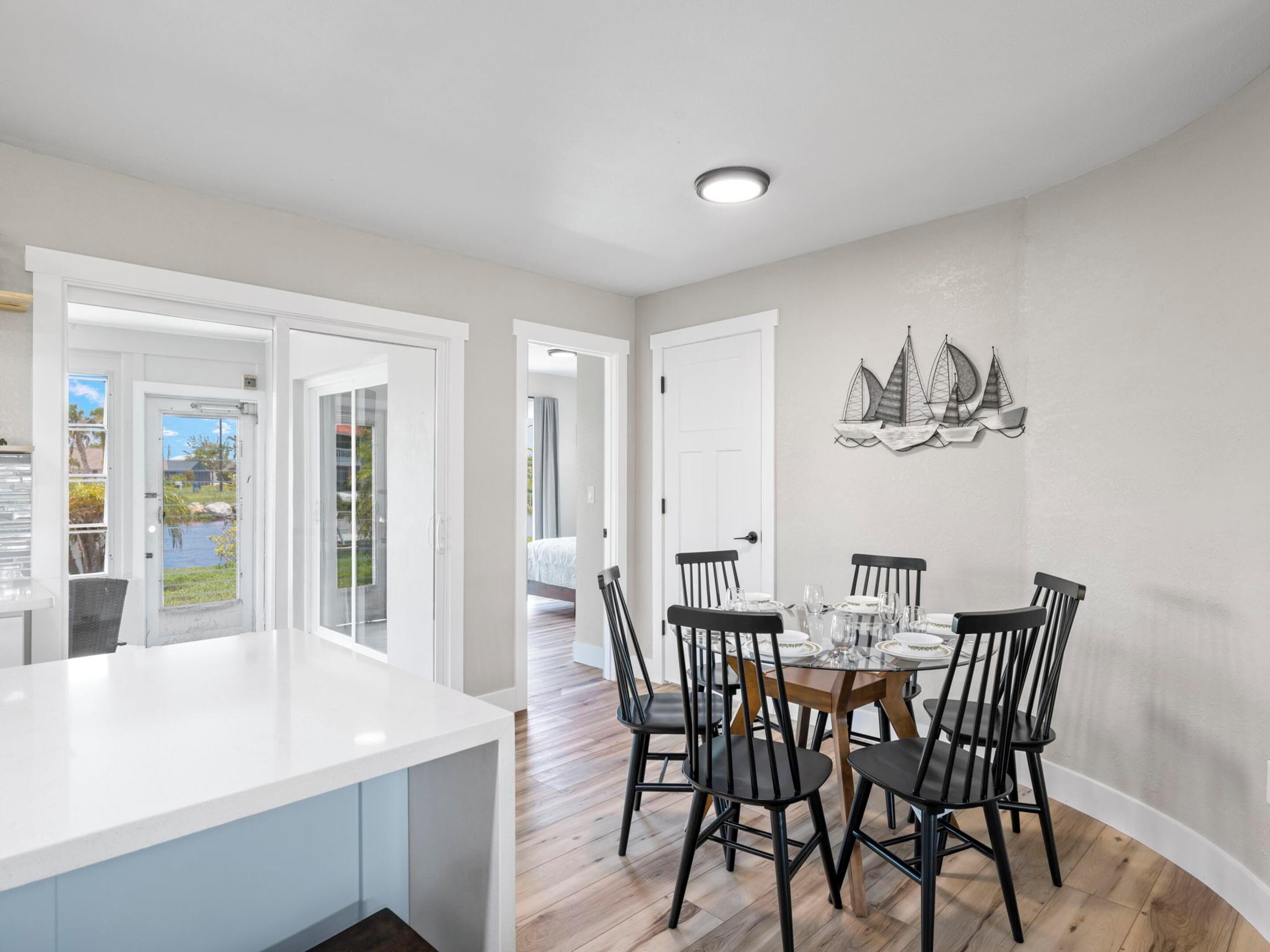 A modern kitchen with a central island, dining area with six chairs, and a view through glass doors to an outdoor area.