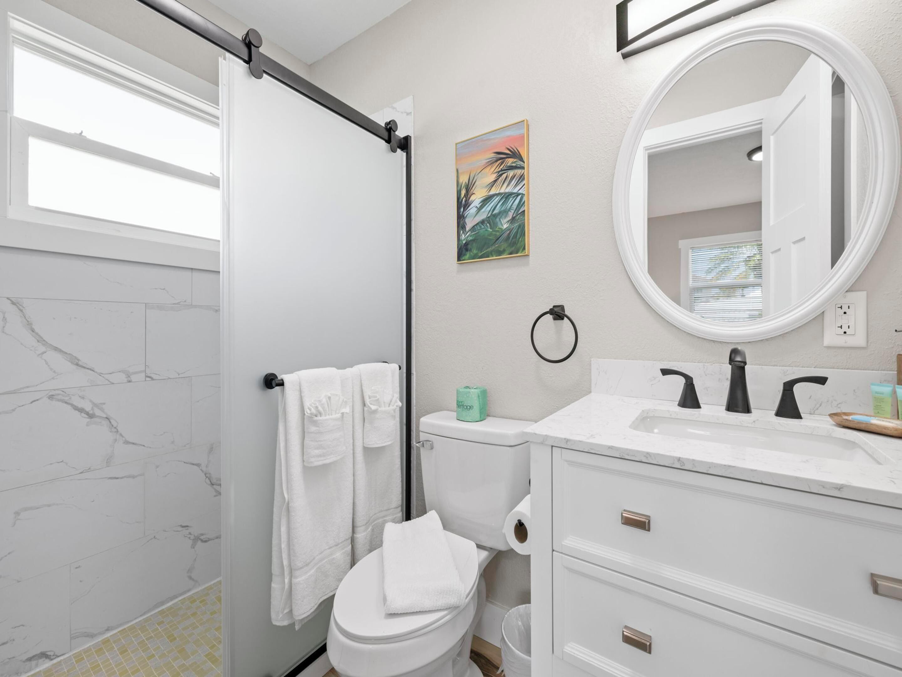 A bathroom with a white toilet, a white sink with a marble countertop, a shower with a black towel rack, and a painting on the wall.