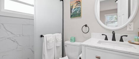 A bathroom with a white toilet, a white sink with a marble countertop, a shower with a black towel rack, and a painting on the wall.