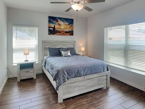 A bedroom with a bed, two bedside tables, a ceiling fan, and a large window with blinds. - Breezy Villa by Distinctive Beach Rentals (Palm Coast)