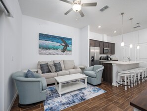 A modern living room with a sofa, armchairs, a coffee table, and a kitchen area with a refrigerator, microwave, and bar stools. - Breezy Villa by Distinctive Beach Rentals (Palm Coast)
