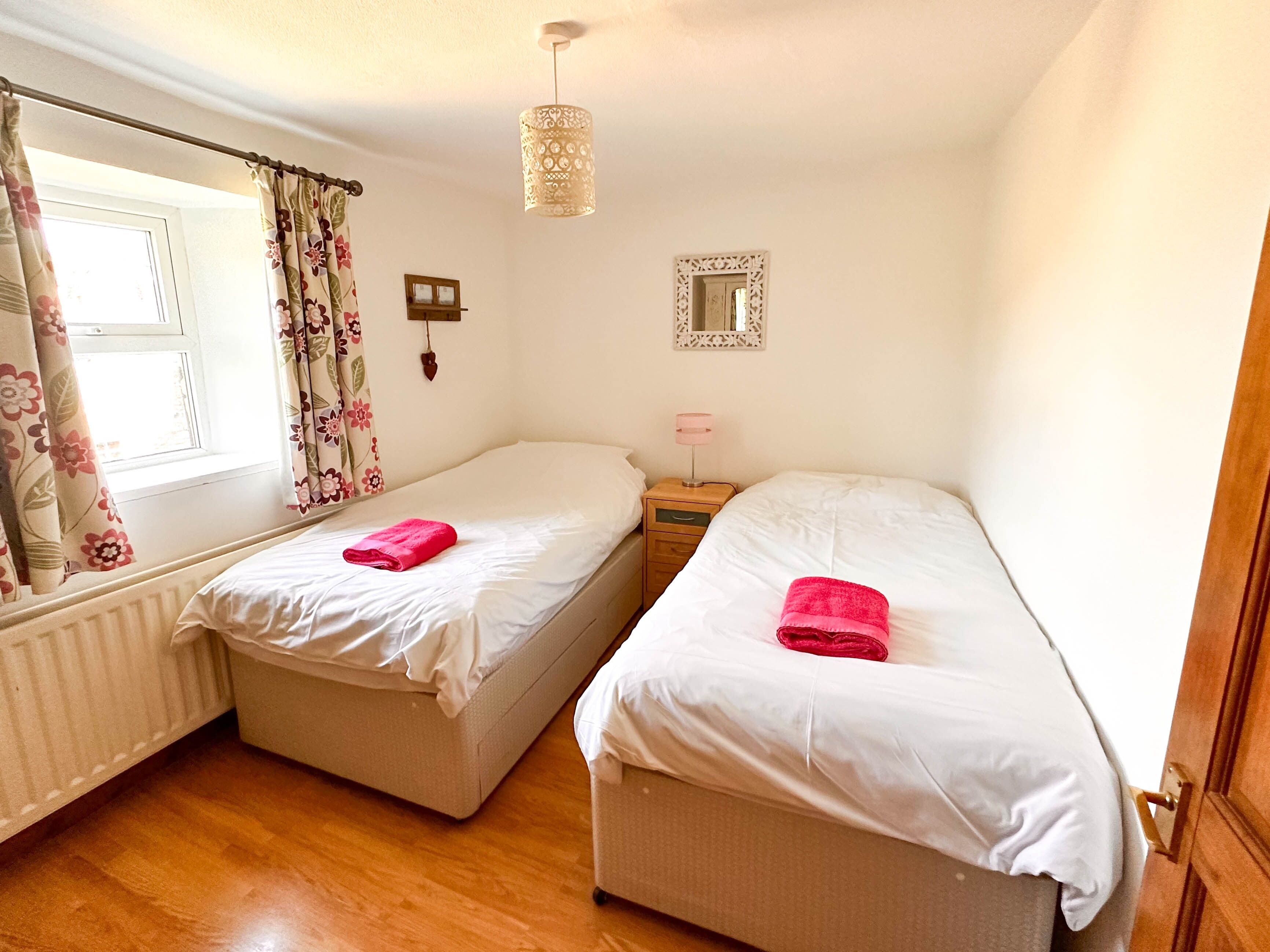 A bedroom with two single beds, a wooden floor, a window with floral curtains, a bedside lamp, and a mirror on the wall.