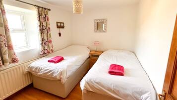 A bedroom with two single beds, a wooden floor, a window with floral curtains, a bedside lamp, and a mirror on the wall.
