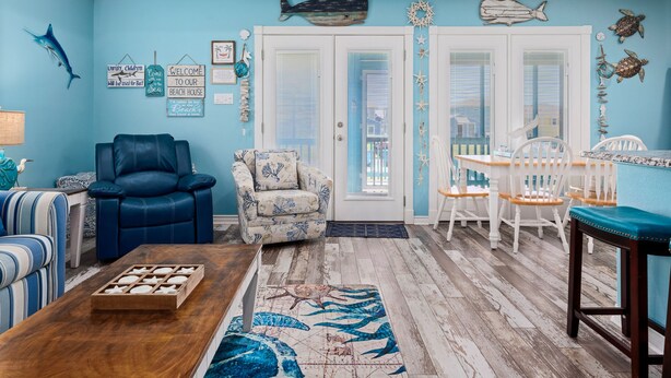 A beach-themed living room with a blue wall, a blue sofa, a patterned armchair, a wooden coffee table, and a dining area with white chairs and a blue tablecloth. - Turtle's Nest by Miss Kitty’s (Rockport)