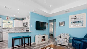 A modern living room with a kitchen area, featuring a wooden coffee table, a blue sofa, and a television mounted on the wall. - Turtle's Nest by Miss Kitty’s (Rockport)