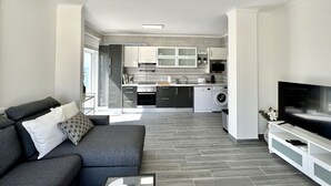 Un appartamento moderno con un divano grigio, una TV a schermo piatto e una cucina con mobili e elettrodomestici bianchi.