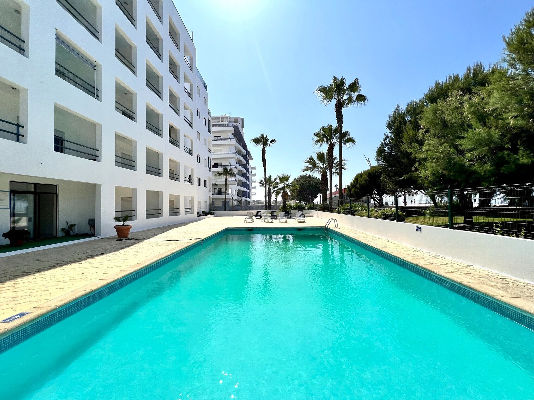 Una piscina con agua azul cristalina, rodeada por un edificio blanco con balcones y un área cercada con palmeras.