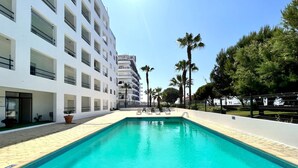 Una piscina con acqua cristallina e di colore blu, circondata da un edificio bianco con balconi e un'area recintata con palme.