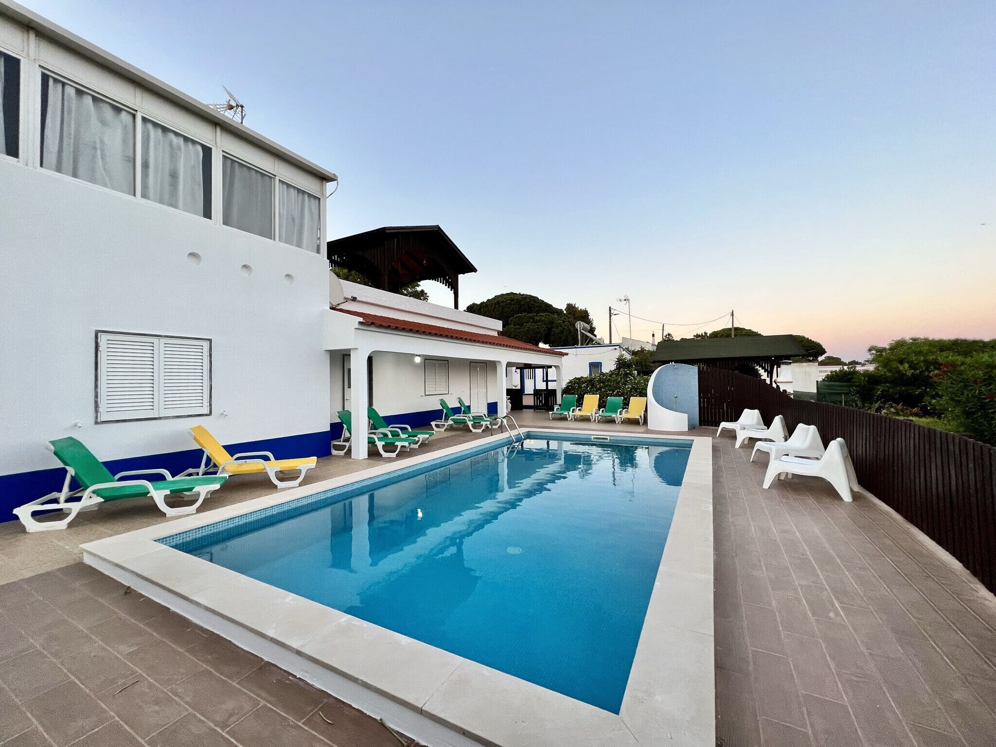 A swimming pool with lounge chairs and a covered area, surrounded by a building with a tiled roof and a fenced garden.