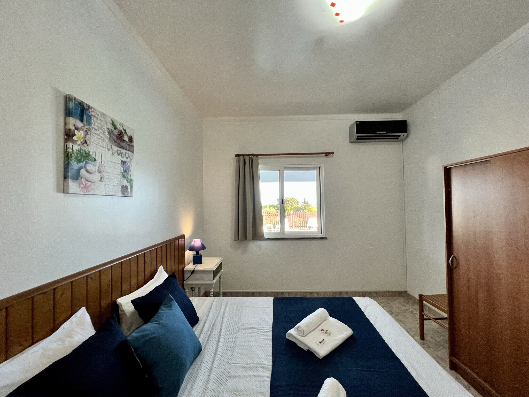 Een slaapkamer met een bed, nachtkastje, lamp, raam, airconditioning en een deur.